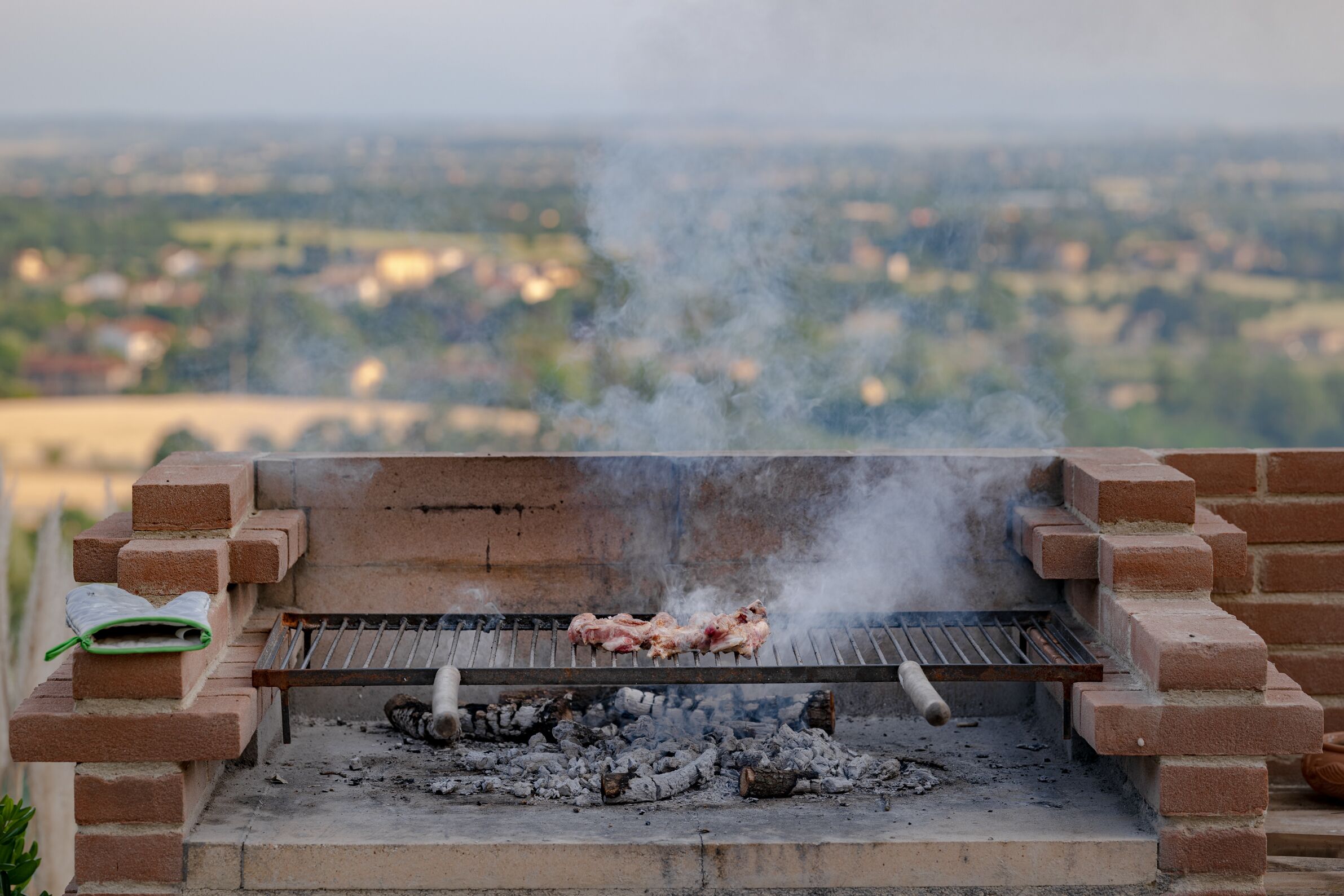 A barbecue grill with meat cooking, surrounded by a brick structure, and a view of a distant landscape.