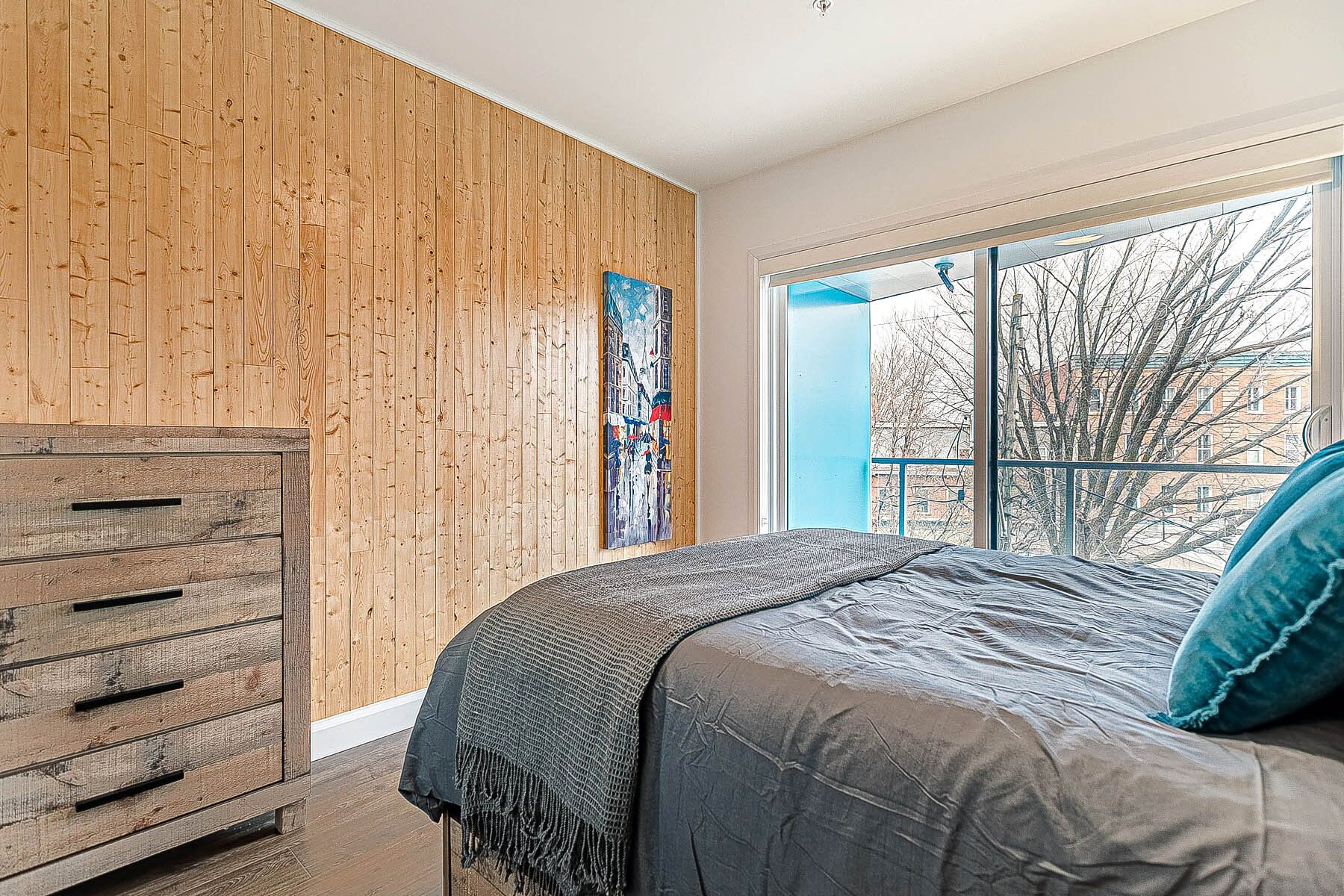 Un dormitorio con pared de madera, una cama con manta gris y una ventana con vistas a edificios.