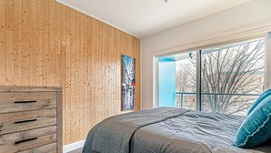 A bedroom with a wooden wall, a bed with a grey blanket, and a window with a view of buildings. - The Kaméléon, The hut (Québec City)