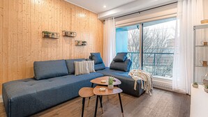 A modern living room with a blue sofa, a small wooden coffee table, and a balcony with a view of trees and buildings. - The Kaméléon, The hut (Québec City)