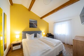 A hotel room with a large bed, two bedside tables with lamps, a gray headboard, a painting on the wall, a window with sheer curtains, and a wooden cabinet.