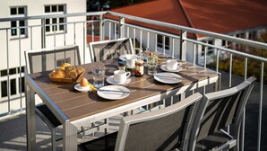 Una mesa de comedor dispuesta en un balcón, con sillas, platos, tazas y una cesta de pan.