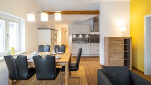 Una cocina moderna con armarios blancos, una mesa de comedor de madera con sillas negras y un sofá gris en una habitación luminosa con paredes amarillas.