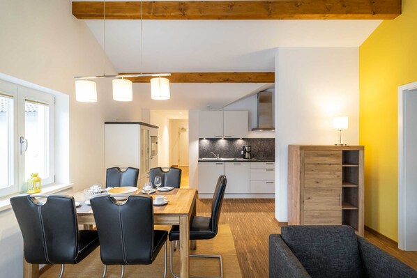 A modern kitchen with white cabinets, a wooden dining table with black chairs, and a grey sofa in a bright room with yellow walls.