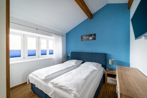 A bedroom with two beds, a blue wall, a wooden ceiling, a bedside table with a lamp, and a window with blinds.