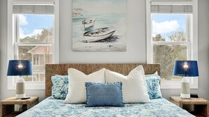 Een slaapkamer met een bed, twee bedlampen, een schilderij van een boot en een raam met jaloezieën.
