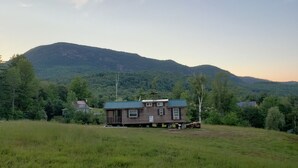 Una caba ña con terraza, rodeada por un campo cubierto de hierba y una montaña boscosa al fondo.