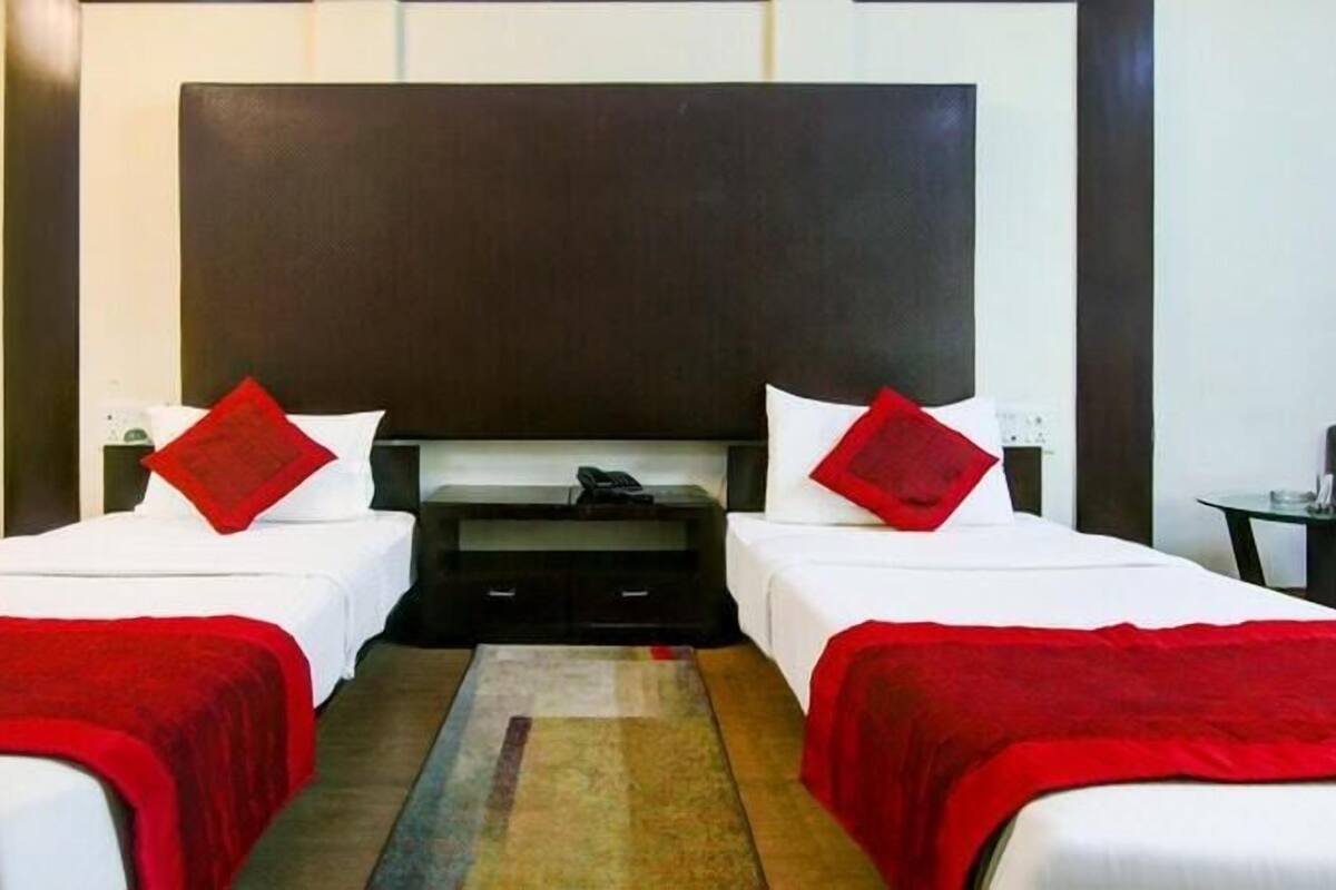 Quarto de hotel com duas camas, cada uma com manta e almofadas vermelhas, uma cabeceira escura grande e uma mesa pequena com abajur.
