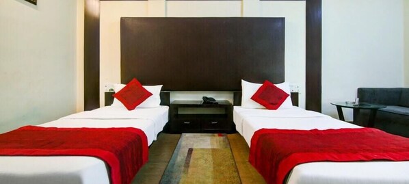 A hotel room with two beds, each with a red throw blanket and pillows, a large dark headboard, and a small table with a lamp. - Liveinn Style (Gurugram)
