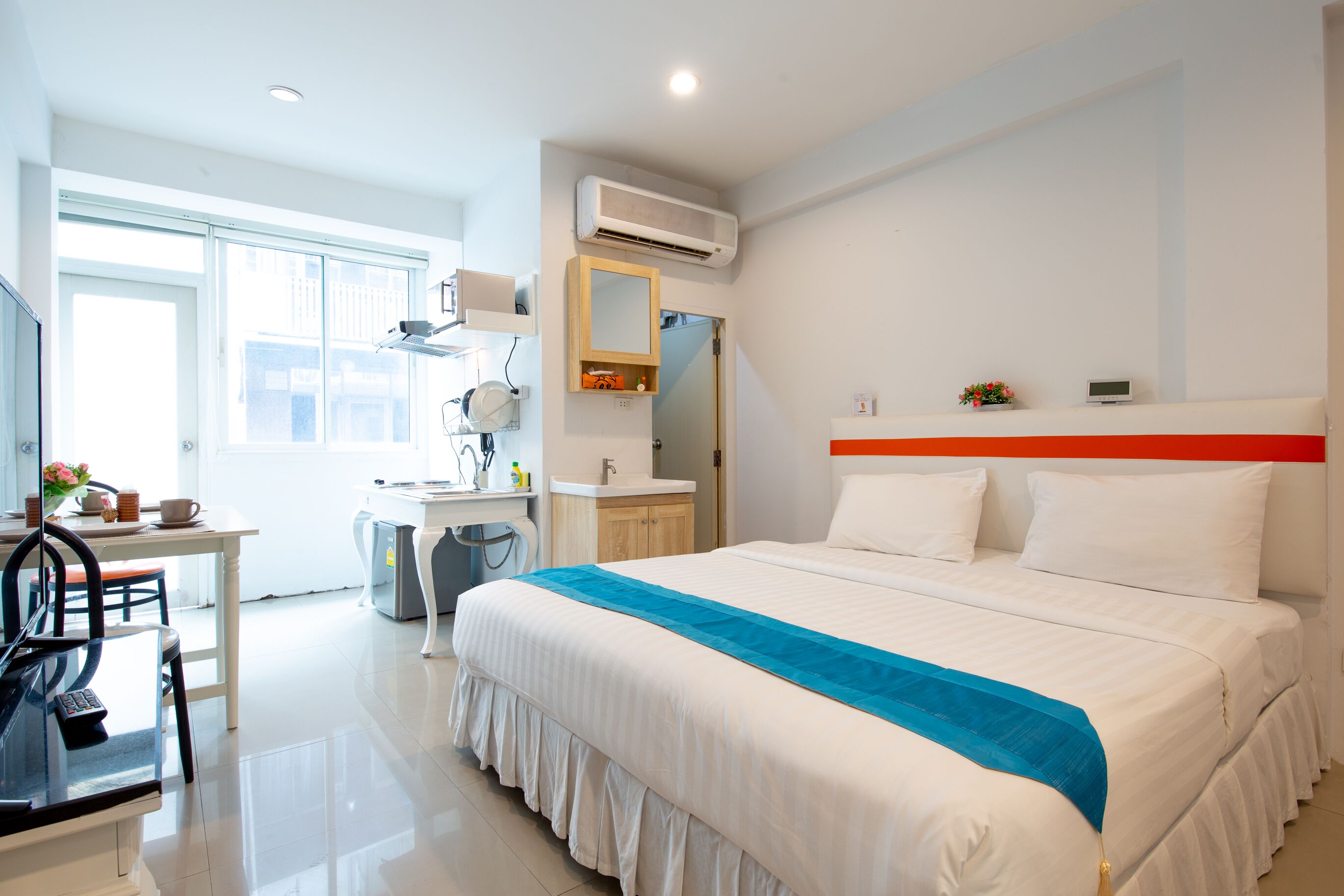 A hotel room with a bed, a desk, a chair, a mirror, a sink, and an air conditioning unit.