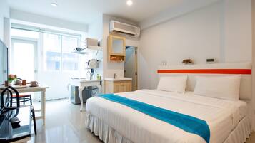 A hotel room with a bed, a desk, a chair, a mirror, a sink, and an air conditioning unit.