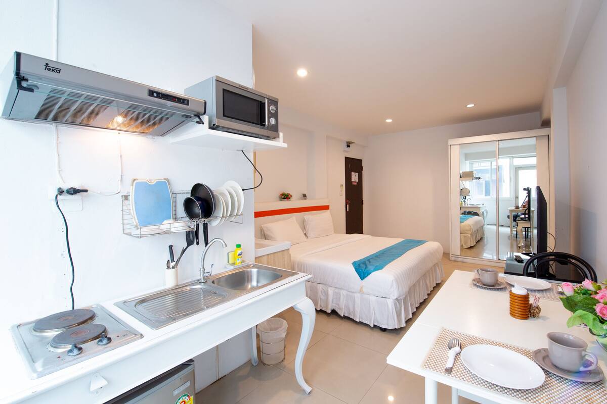 Una habitación de hotel moderna con kitchenette, una cama con ropa de cama blanca, una mesa de comedor con posavasos y un espejo que refleja la habitación.