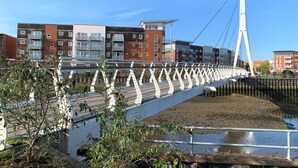 Un puente peatonal con barandilla blanca y diseño atirantado, que cruza un cuerpo de agua con edificios al fondo.