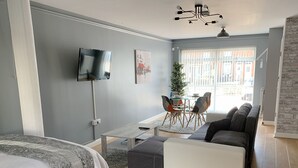 Una habitación moderna y luminosa con una cama, un sofá, una pequeña mesa de comedor con sillas, un televisor y un amplio ventanal con persianas.