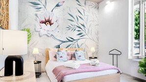 Un dormitorio con un amplio mural floral en la pared, una cama con manta rosa, dos mesitas de noche con lámparas y una ventana con vistas a zonas verdes.