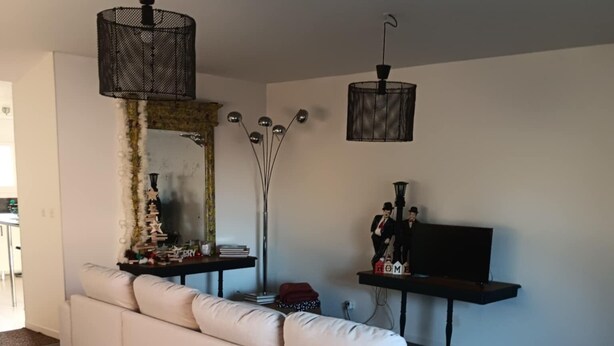 Uma sala de estar moderna com um sofá branco, uma televisão sobre um móvel e objetos decorativos em uma mesa lateral.