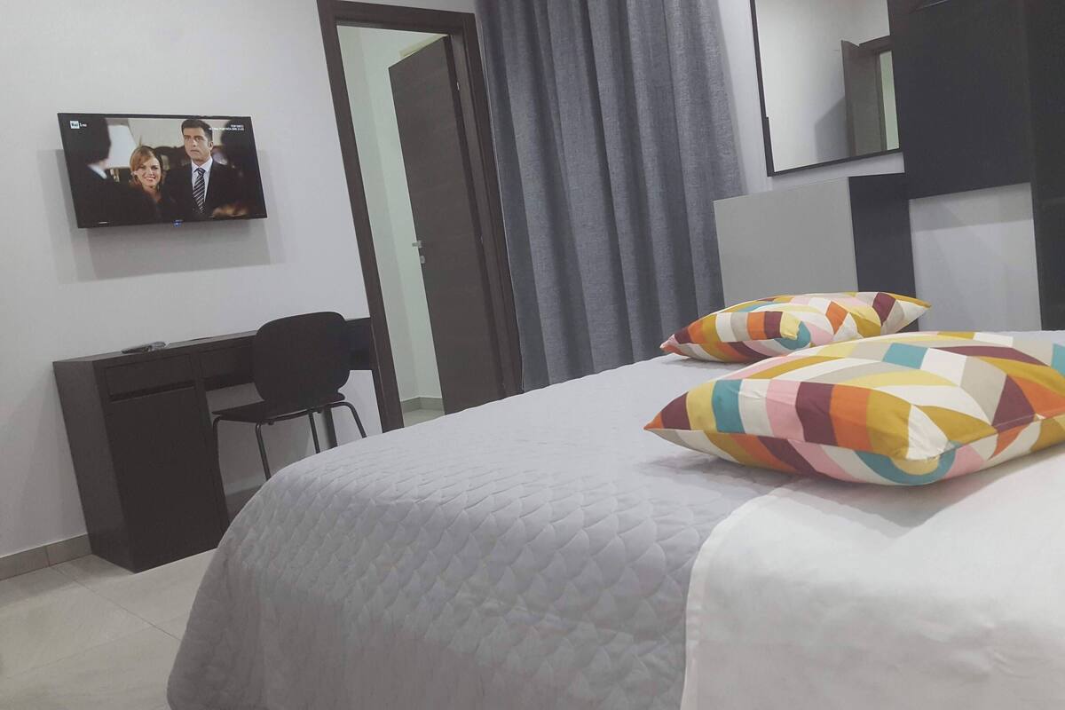 Ein Hotelzimmer mit Bett, zwei farbenfrohen Kissen, einem Schreibtisch, einem Stuhl, einem Fernseher und einem Spiegel.