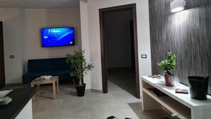 A modern living room with a flat-screen TV, a blue sofa, a coffee table, two potted plants, and a white console table with a black bucket. - Idilliaco Inn (Motta Sant'Anastasia)