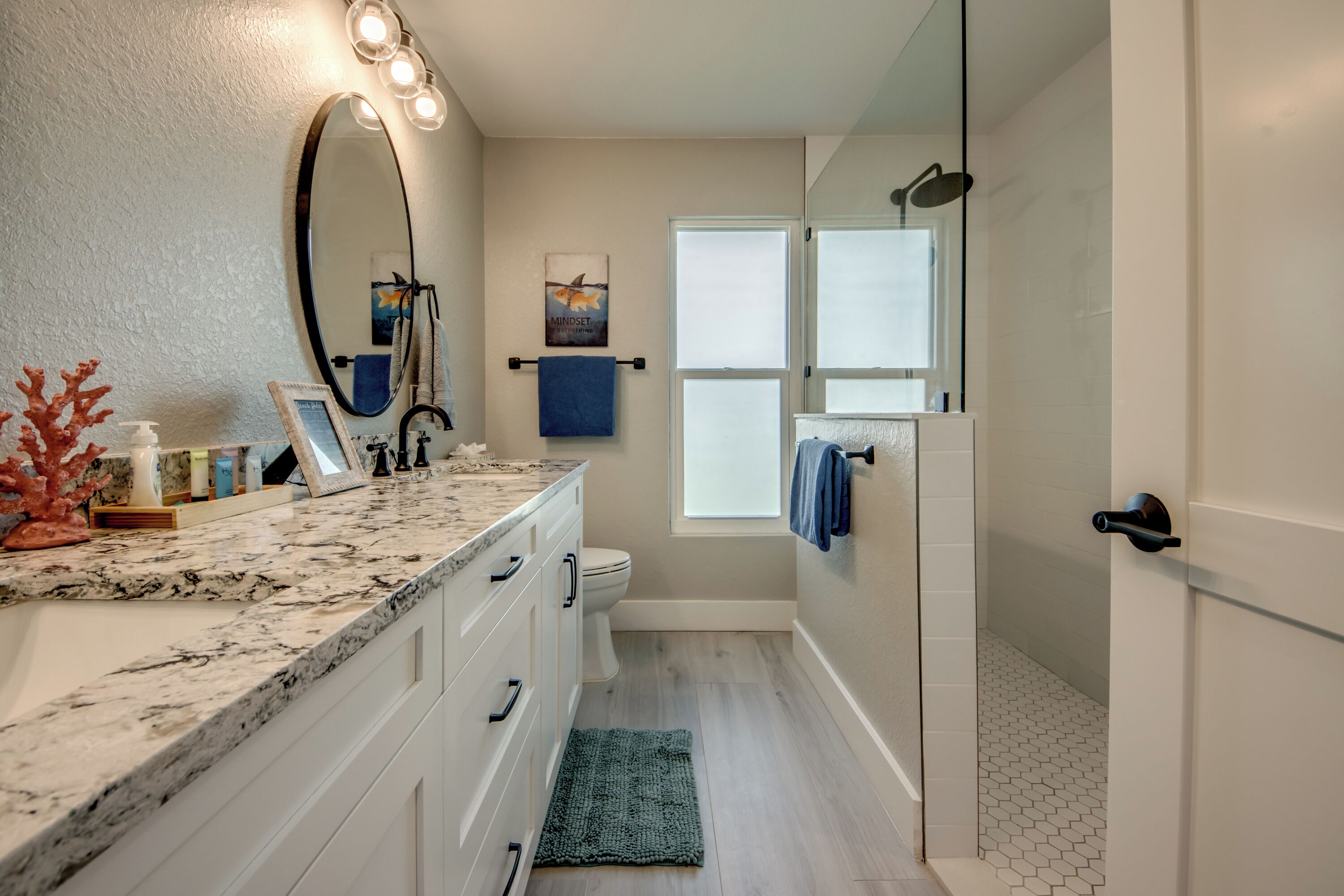 A bathroom with a marble countertop, a large mirror, a shower with a glass door, and a window.