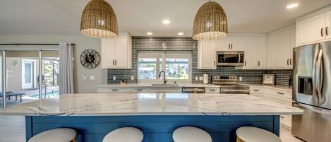 A modern kitchen with a large island, white cabinets, stainless steel appliances, and wicker pendant lights.