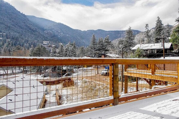 A view from a balcony with a metal railing, overlooking a snowy landscape with mountains, trees, and a road with vehicles.