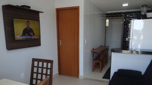 A living room with a TV mounted on the wall, a wooden dining table with chairs, a black sofa, and a kitchen area with a refrigerator and tiled backsplash.