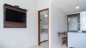 A modern room with a flat-screen TV mounted on the wall, a wooden-framed door leading to a bedroom, a small kitchen area with a refrigerator, and a seating area with a sofa.