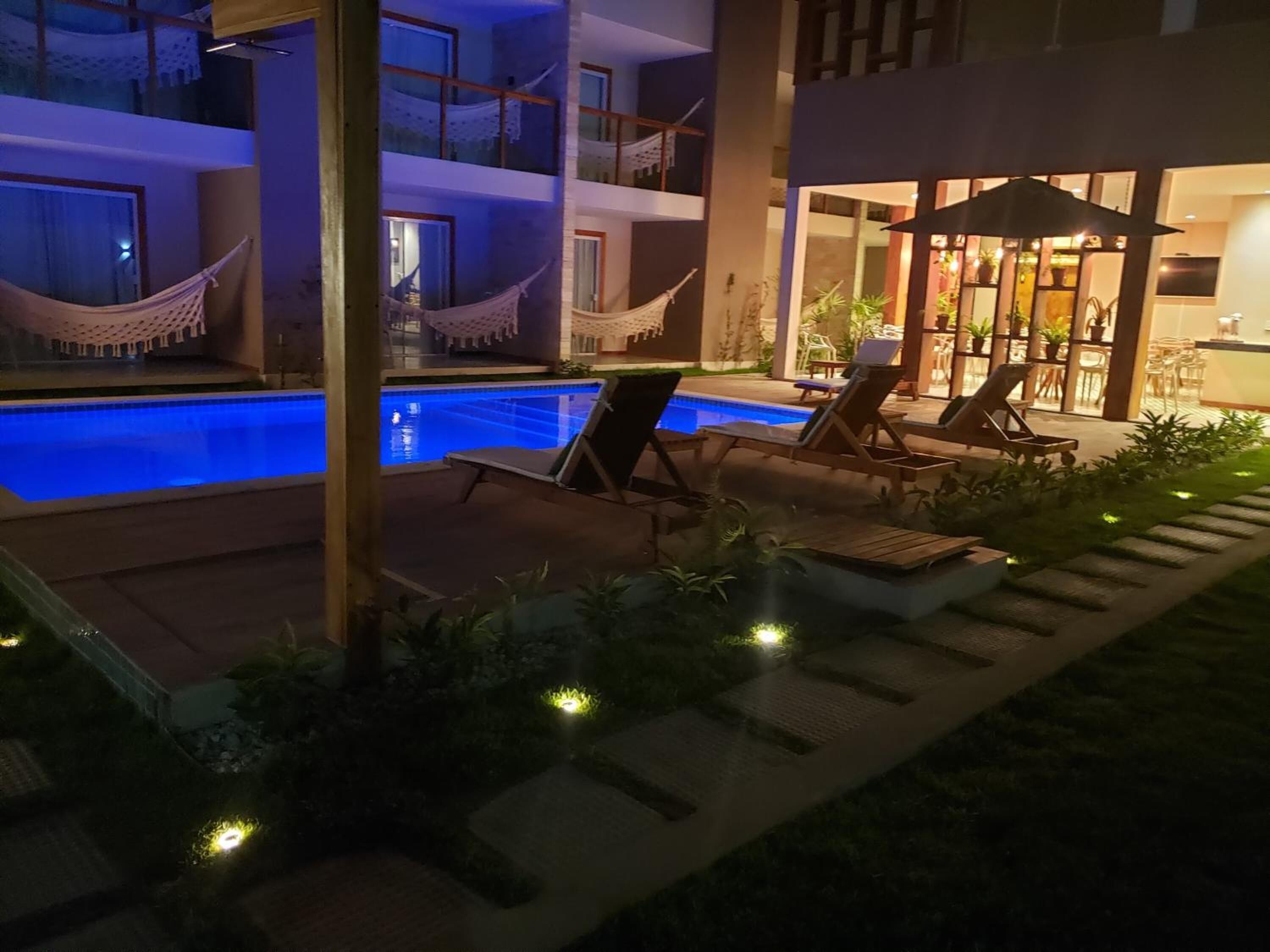 Un área de piscina con sillones reclinables y hamacas, rodeada por un edificio de varios pisos y grandes ventanales.