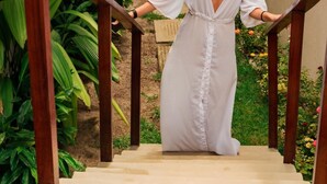 Mulher descendo uma escadaria de madeira, vestindo um vestido branco, rodeada por vegetação e um muro de pedra.