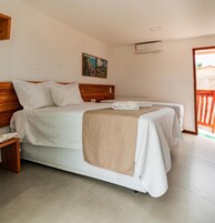 Cama doble con cabecera de madera, manta beige y ropa de cama blanca. Se observa una mesita de noche con lámpara, aire acondicionado instalado en la pared y un cuadro sobre la cama.