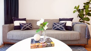 Een woonkamer met een grijze bank, twee decoratieve kussens, een vaas met bloemen, een boek en een plant.