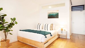 Een moderne slaapkamer met een houten bedframe, witte beddengoed, een groene plaid en een klein nachtkastje met een lamp.