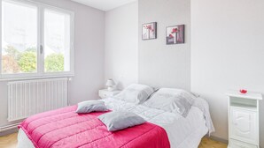 Ein Schlafzimmer mit einem Bett, weißen Nachttischen, einer weißen Kommode und einem Fenster mit Blick auf Grünflächen.