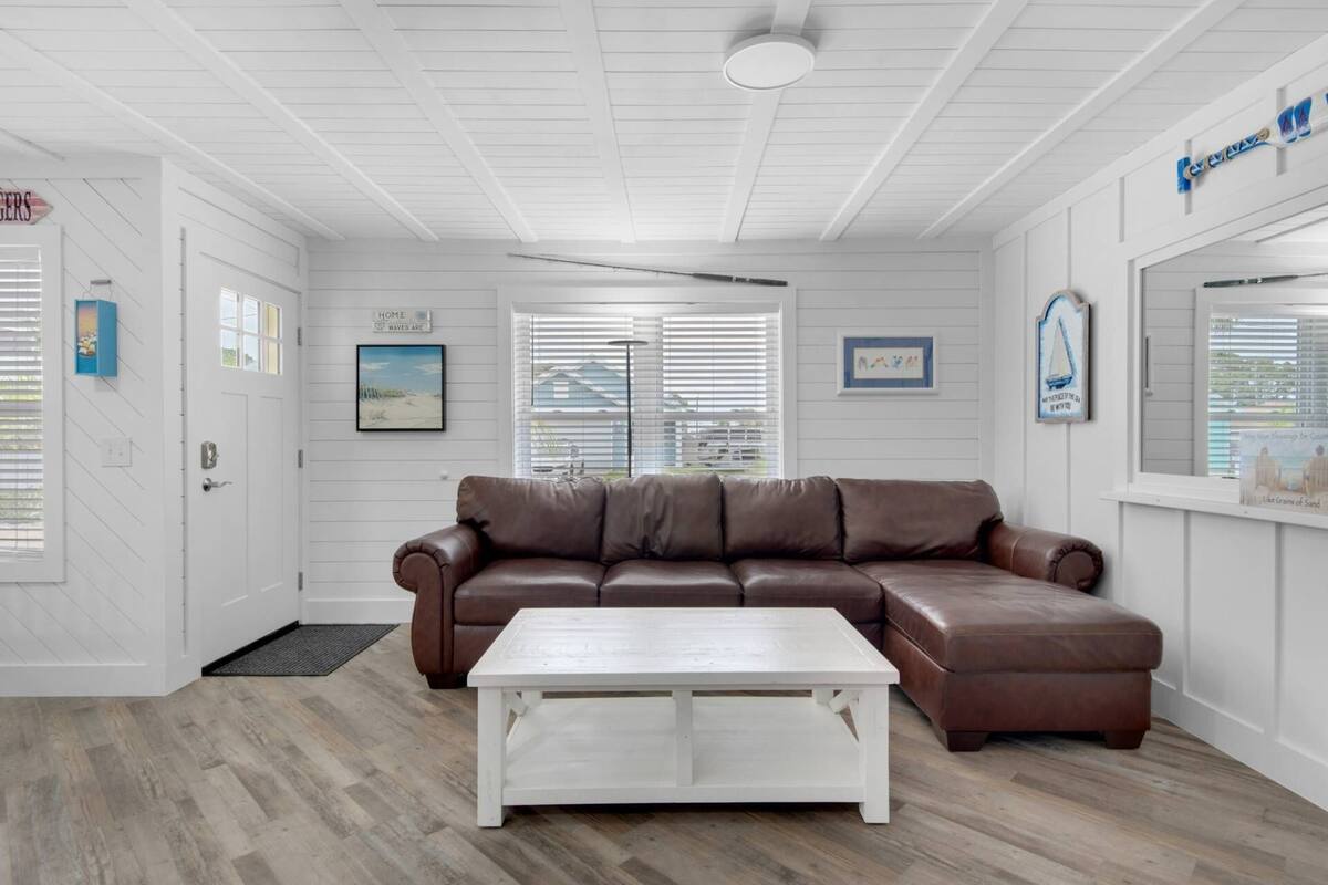 Ein Wohnzimmer mit braunem Ledersofa, einem weißen Couchtisch und einer weißen Tür mit kleinem Fenster. An den Wänden befinden sich Dekorationen und es gibt einen Deckenventilator.
