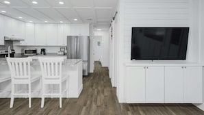 A modern kitchen with white cabinetry, a built-in microwave, a stainless steel refrigerator, and a large flat-screen TV mounted on the wall. - Sunset Escapes B (Panama City Beach)