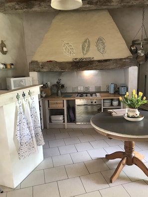 A kitchen with a gas stove, microwave, and oven. There is a hanging pot rack with aprons and a potted plant on the table.
