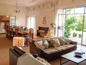 A spacious living room with a large sofa, a dining area with wooden chairs, and a view of greenery outside.