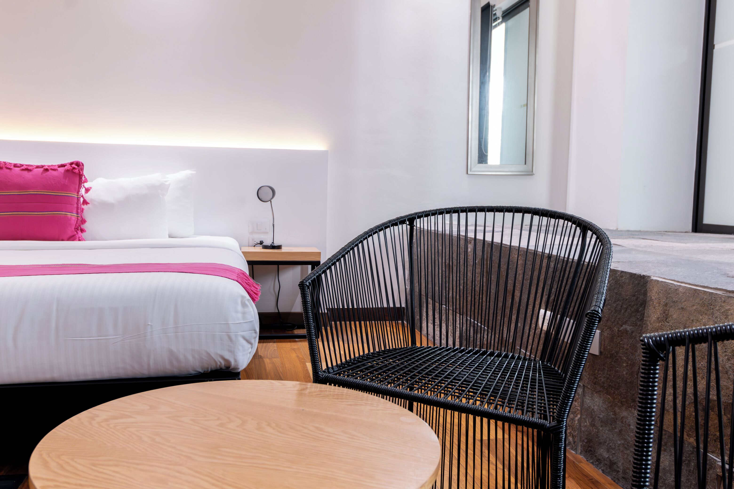A hotel room with a bed, a round wooden table, a black wicker chair, and a nightstand with a lamp.
