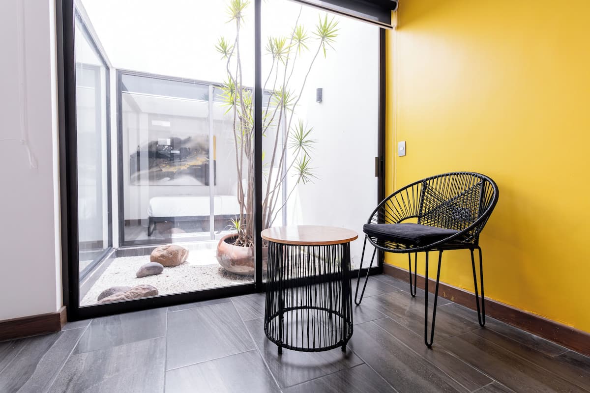 A modern room with a glass door leading to a balcony, a round wooden table, a black wireframe chair, and a yellow wall.