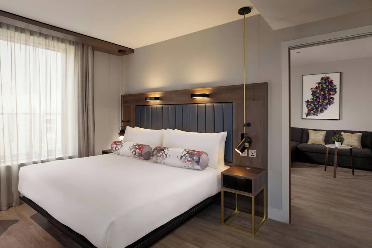 A modern hotel room with a large bed, a bedside table, a lamp, and a view of the living area.