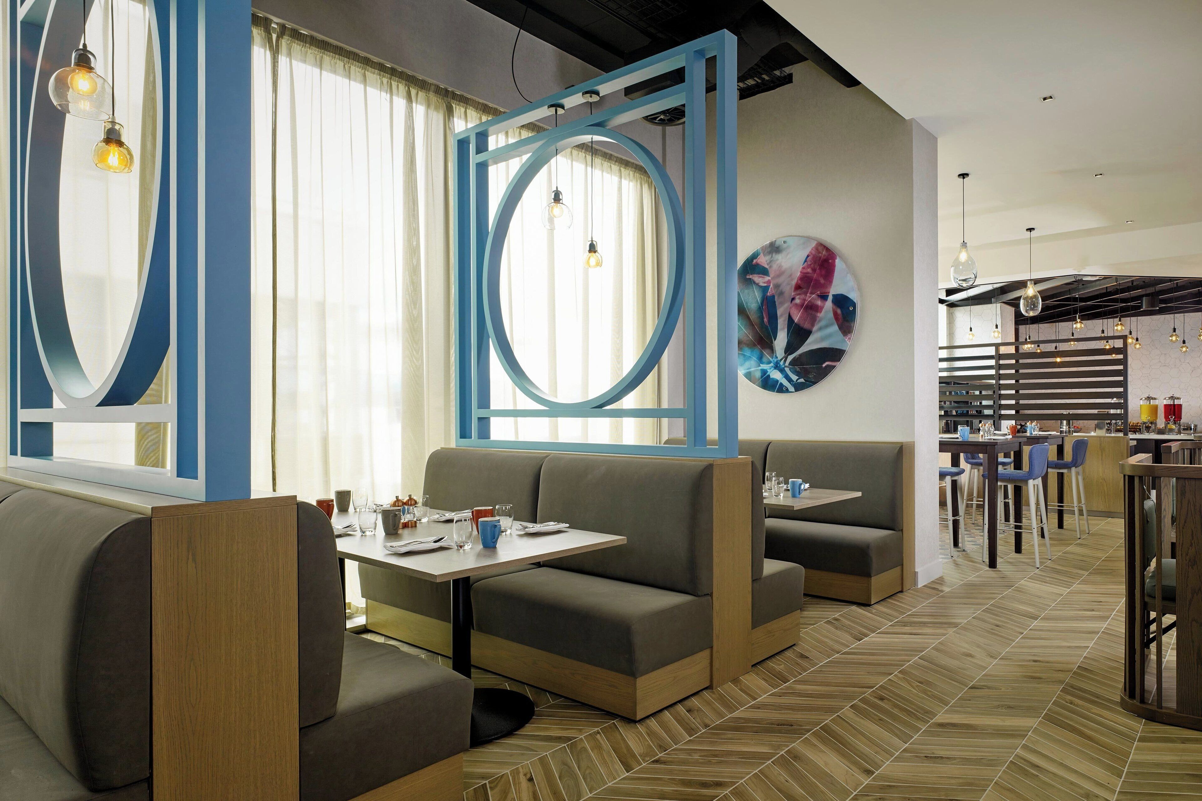 A modern restaurant with booths and tables, a large window with curtains, and a decorative wall piece.