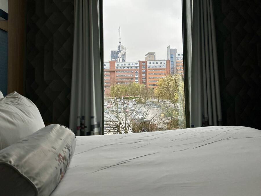 A hotel room with a bed, a window with curtains, and a view of a cityscape.