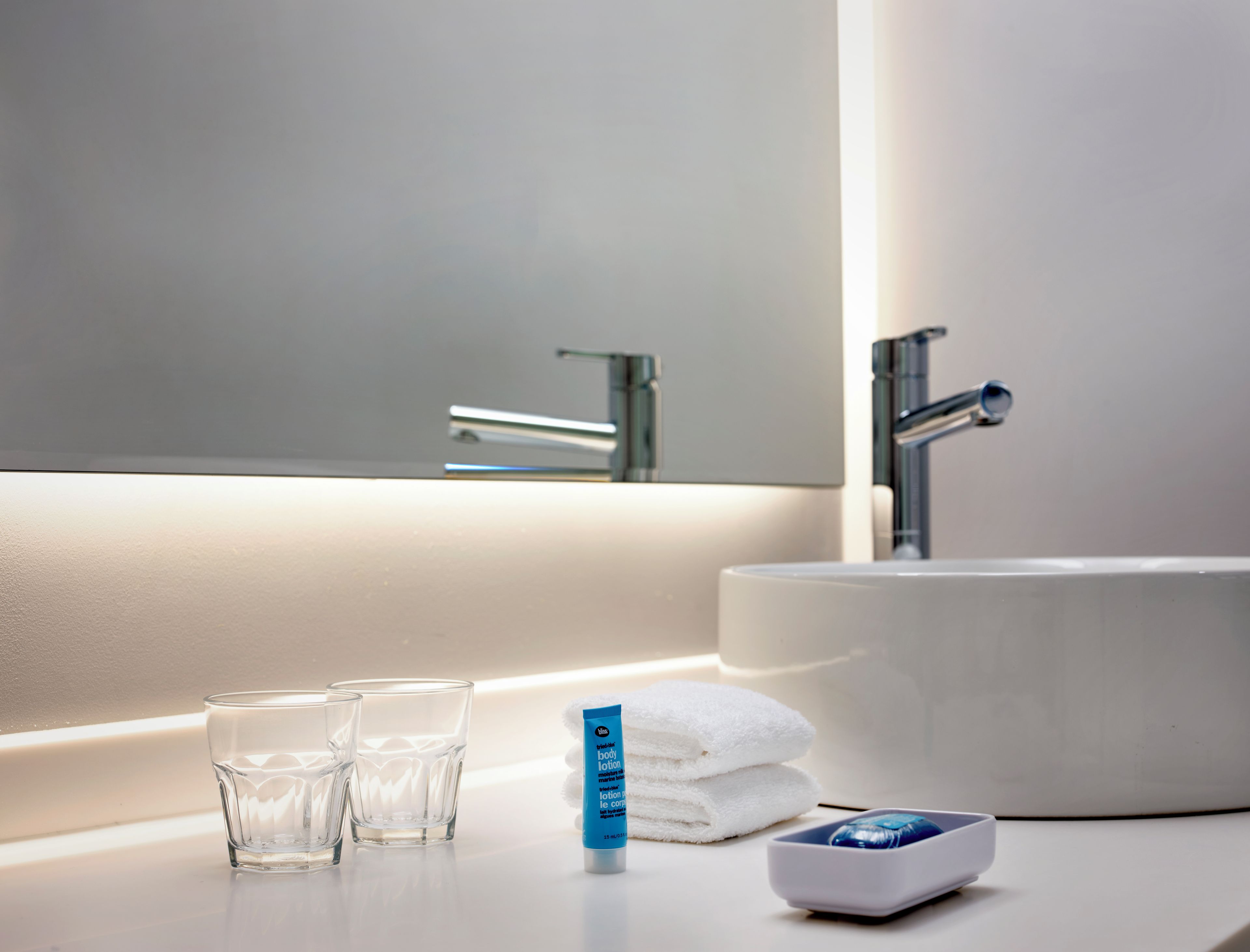 Un baño con un lavabo blanco, dos vasos, un tubo de crema corporal, una toalla y un jabonero.