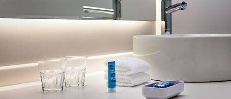 Un baño con un lavabo blanco, dos vasos, un tubo de crema corporal, una toalla y un jabonero.