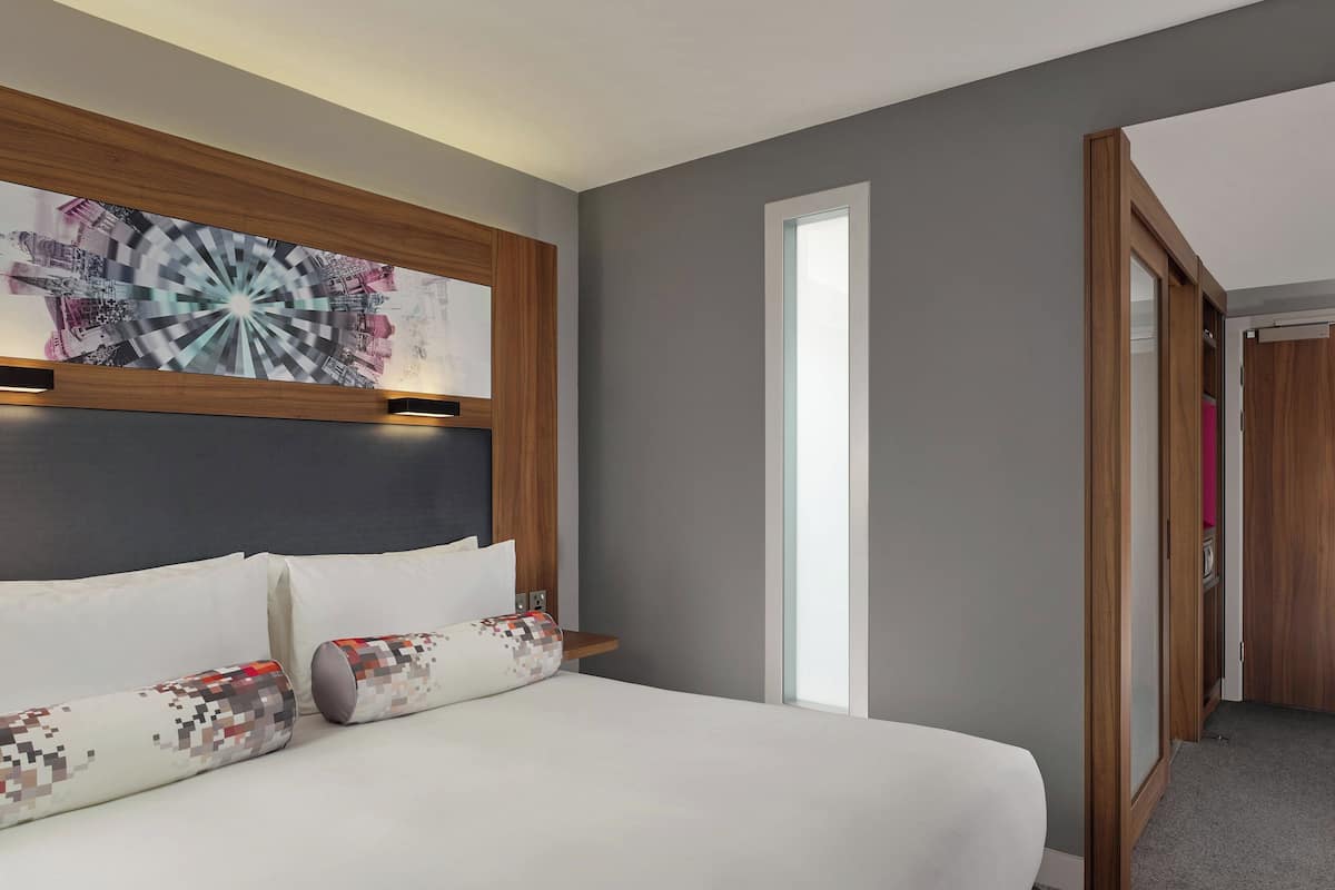 A modern hotel room with a large bed, a headboard with a decorative panel, a bedside table, and a view of a hallway.