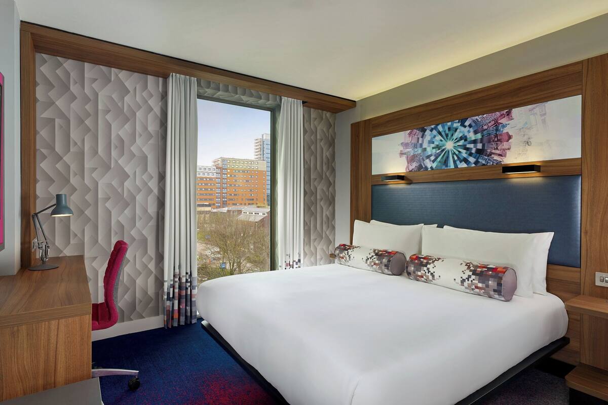 A hotel room with a large bed, a desk with a lamp, a chair, and a view of buildings through the window.