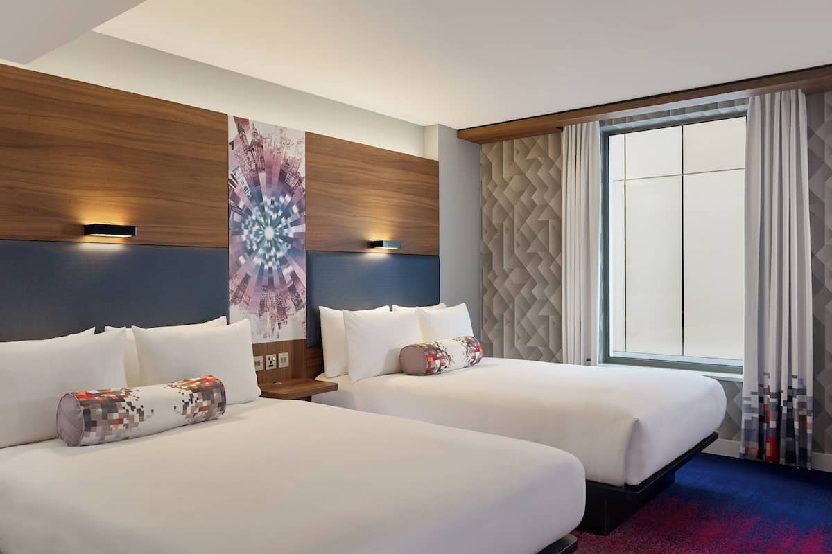 A hotel room with two beds, a large window with curtains, a wall with a geometric pattern, and a wall-mounted light fixture.