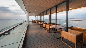 A long, covered outdoor seating area with tables and chairs, overlooking a body of water with boats.