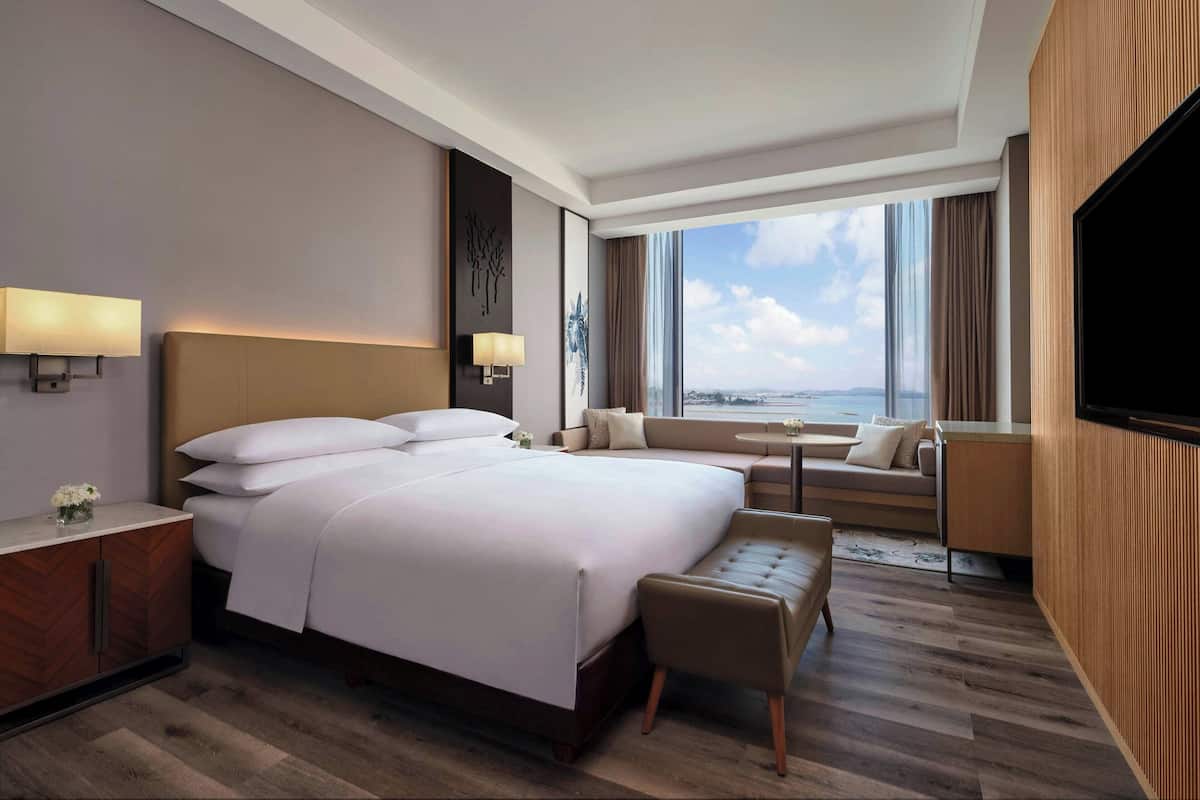 Habitación de hotel moderna con una cama grande, una zona de estar con sofá y sillones, un televisor y vistas a la ciudad.