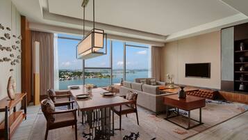 A spacious living room with a dining area, large windows offering a view of a body of water, and a flat-screen TV mounted on the wall.